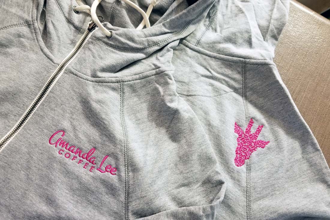 Gray zip-up hoodie sweatshirts with Amanda Lee Coffee embroidered on the left chest and the shape of a giraffe head made out of coffee beans on the back right shoulder.