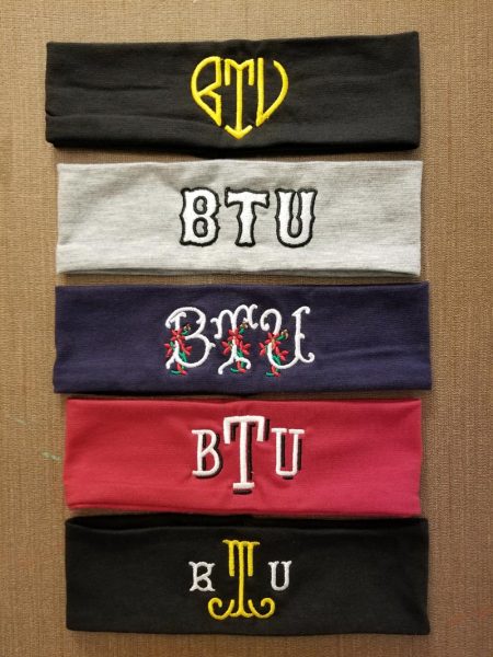 Five wide stretchy fabric headbands: the black one is embroidered with BTU in a heart shape, the gray one with BTU in white letters outlined in black, the navy one with BTU in white letters with red flowers, the red one with BTU written in a monogram style, and the black one with T in gold in the middle and B and U in white on either side.