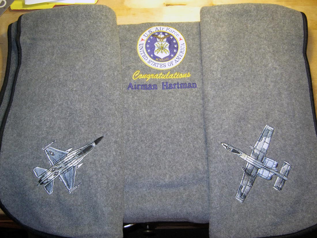 Gray fleece blanket with a military jet embroidered on each of two corners, the US Air Force emblem embroidered on the top center, and the text Congratulations Airman Hartman beneath it.