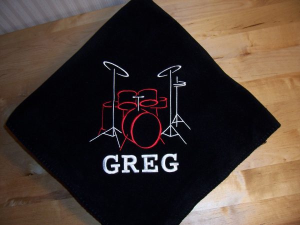 Black fleece blanket embroidered with a drum set design, the drums being red and the snares being white, and the name Greg in white underneath