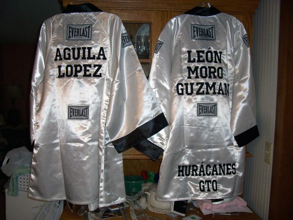 Two shiny white boxing robes with black trim, one embroidered with the name Aguila Lopez on the back, and the other with the name Leon Moro Guzman and the words Huracanes GTO on the back in black.
