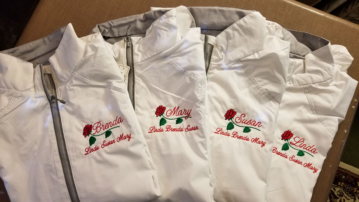 Four white jackets each embroidered on the left chest with a name in red script font, a red rose under the name, and then three other names in smaller red script font under the rose.