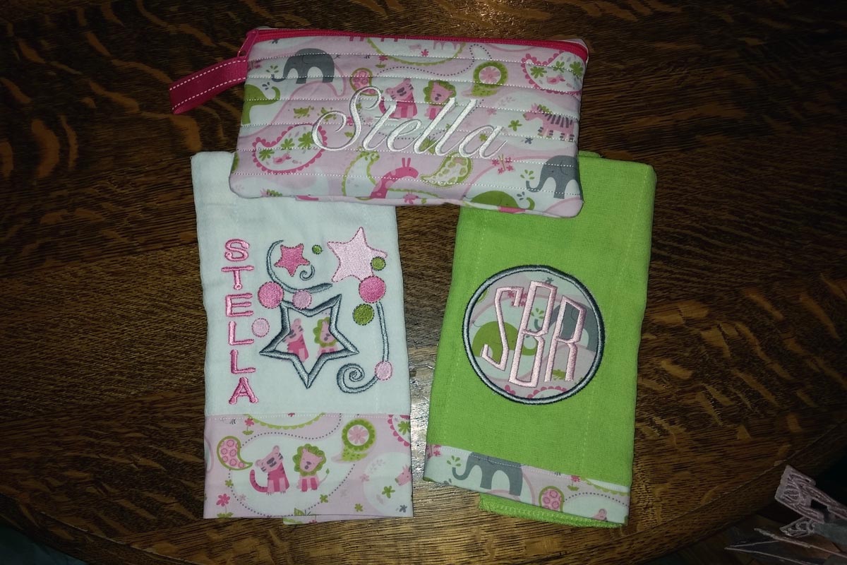 Two baby burp cloths, one embroidered with stars and the name Stella, and one embroidered with the monogram SBR, plus a small handmade zippered pouch also embroidered with the name Stella