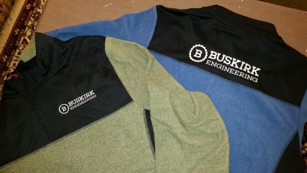 A heathered green and black quarter-zip embroidered with Buskirk Engineering on the left chest laying on top of a sky blue and black quarter-zip with Buskirk Engineering embroidered across the back shoulder area.
