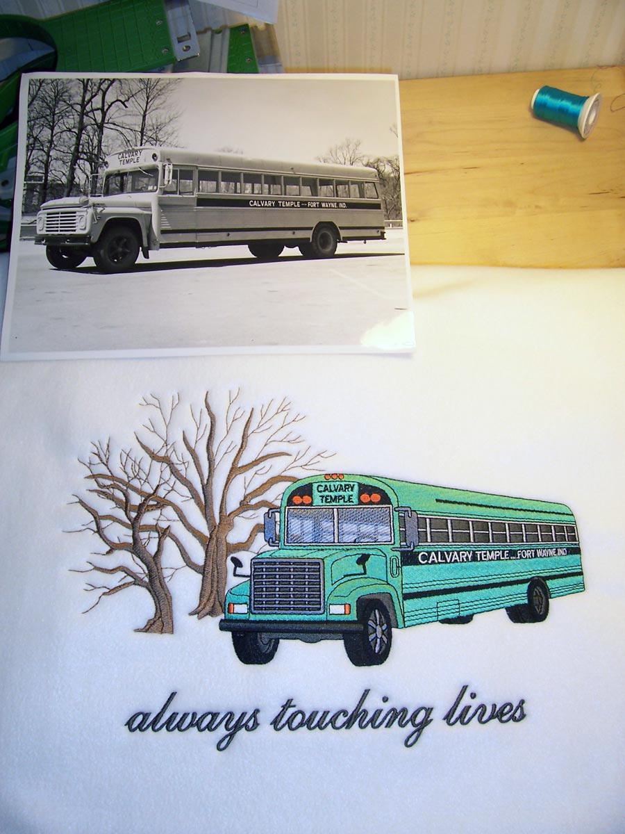 A black and white photograph of a side view of a school bus with the words Calvary Temple Fort Wayne Ind on its side, next to a white casket insert embroidered with an image of that bus next to two bare trees and the words always touching lives underneath.