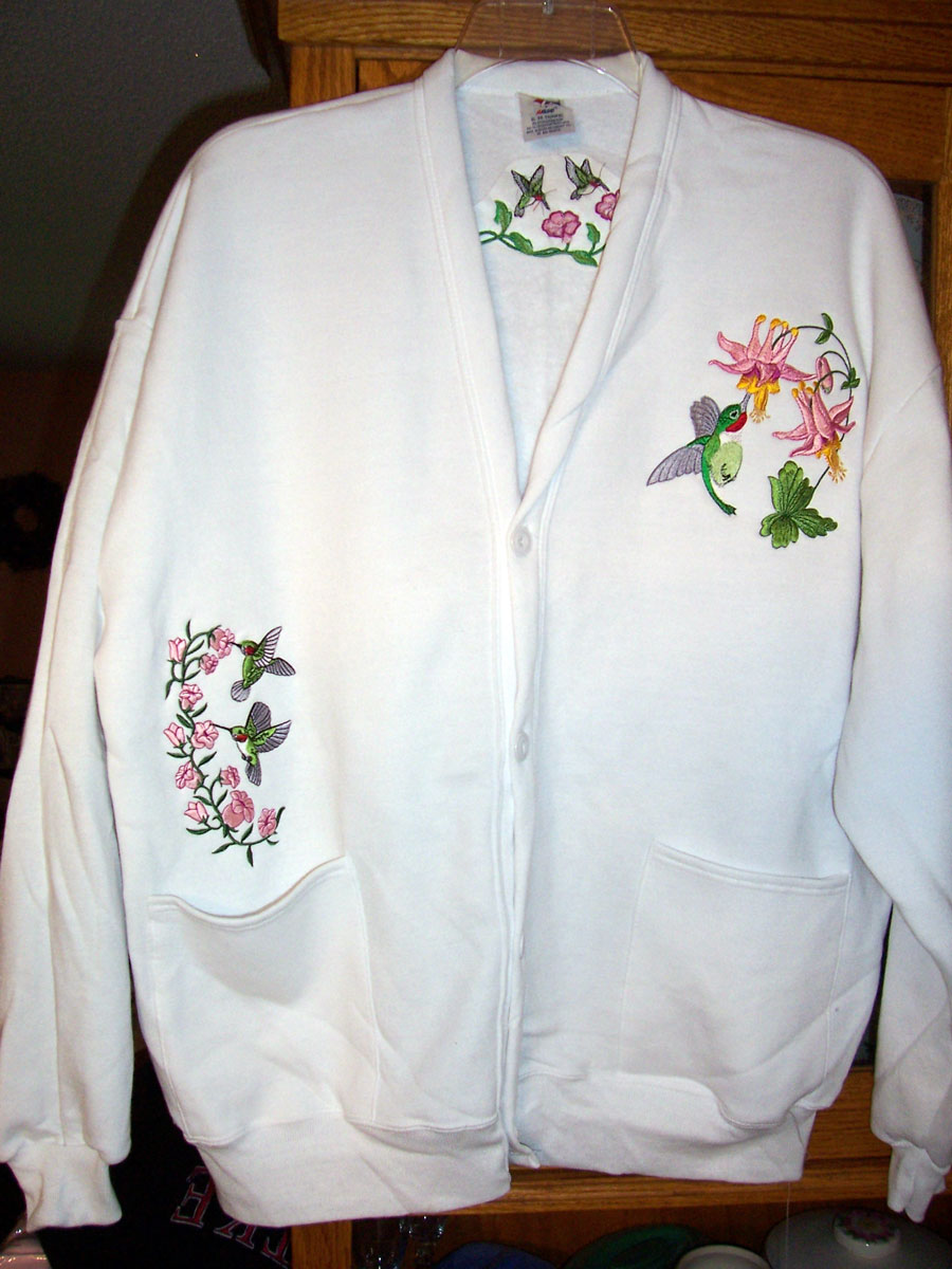 White button-down cardigan embroidered with two hummingbirds and pink flowers above the right pocket, a hummingbird and pink flowers on the left chest, and two hummingbirds and pink flowers on the back upper middle.