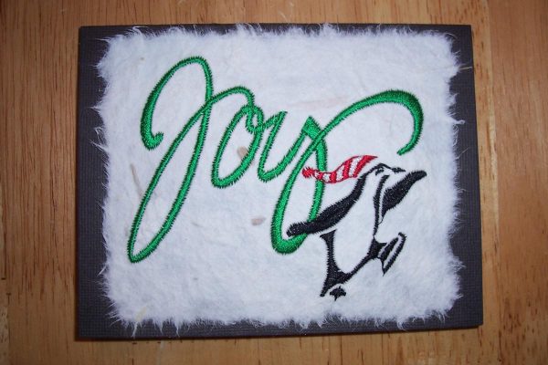 Embroidered Christmas card, the front of which is the word Joy in large green script and an ice skating penguin wearing a red and white knit cap in the lower right corner.