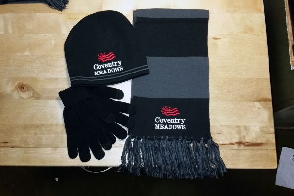 A black knit cap with Coventry Meadows embroidered in white on the front and the Coventry Meadows flag logo embroidered in red above the text sits next to a gray and black fringed scarf with matching embroidery and a set of plain black gloves.