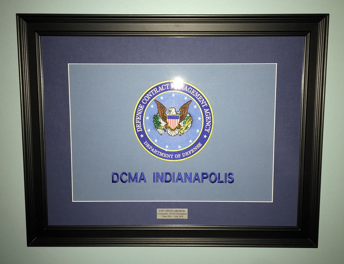 Department of Defense, Defense Contract Management Agency Seal embroidered in blue and gold on a gray background with DCMA Indianapolis embroidered in matching navy blue beneath it, matted and framed.