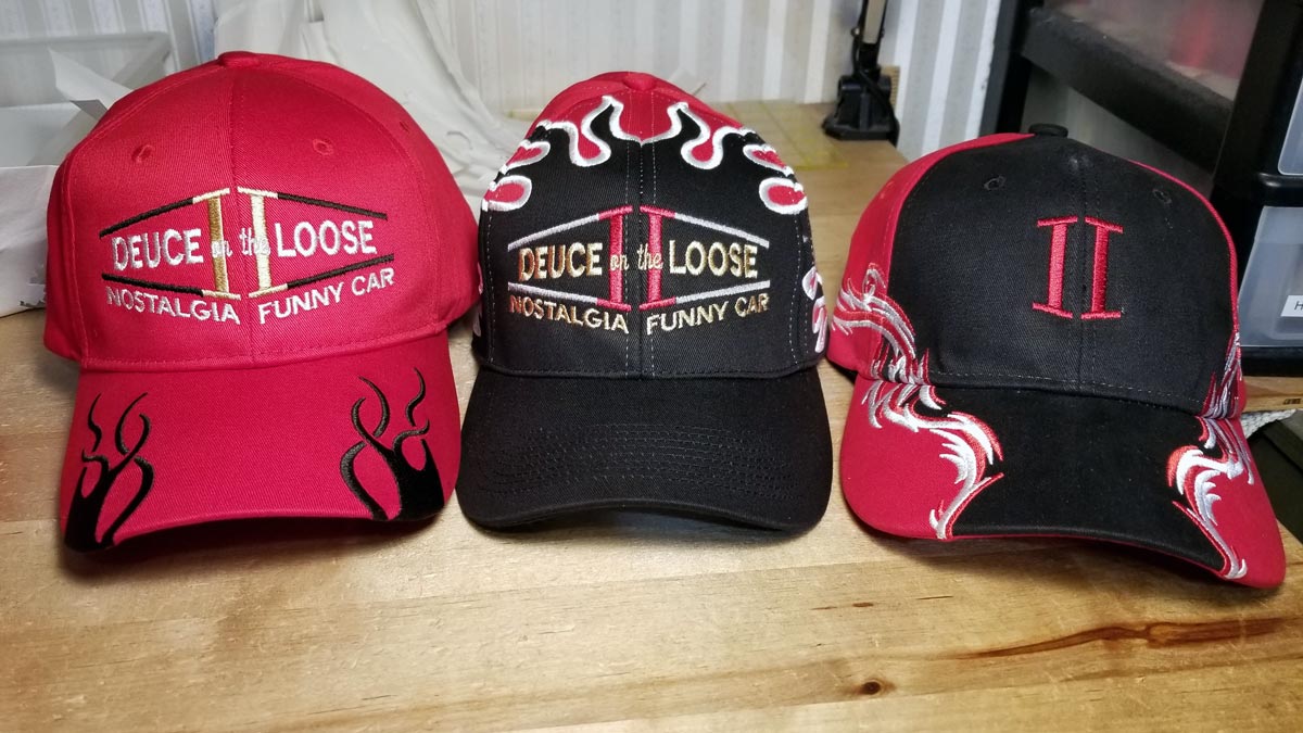 Three baseball caps: the first is red with black flames on the bill and has Deuce on the Loose Nostalgia Funny Car embroidered across the front with the Deuce on the Loose II logo in gold and black behind the text. The second is a red cap with a black bill and black flames on the top and sides, with the same design as the first cap, but the text is gold and the II logo is in red and white. The third cap is red and black with II embroidered in red on the front.