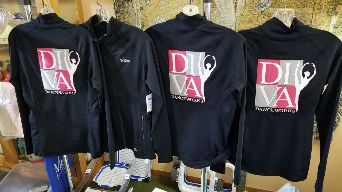 Four black lightweight jackets each with a large pink and gray Diva Danceworks logo embroidered on the back with white lettering. One jacket is turned to the front to show a name embroidered on the left chest.