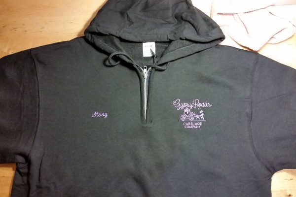 Black or dark gray hoodie with Gypsy Roads Carriage Company logo embroidered in light purple on the left chest with the name Mary embroidered in the same color on the right chest.