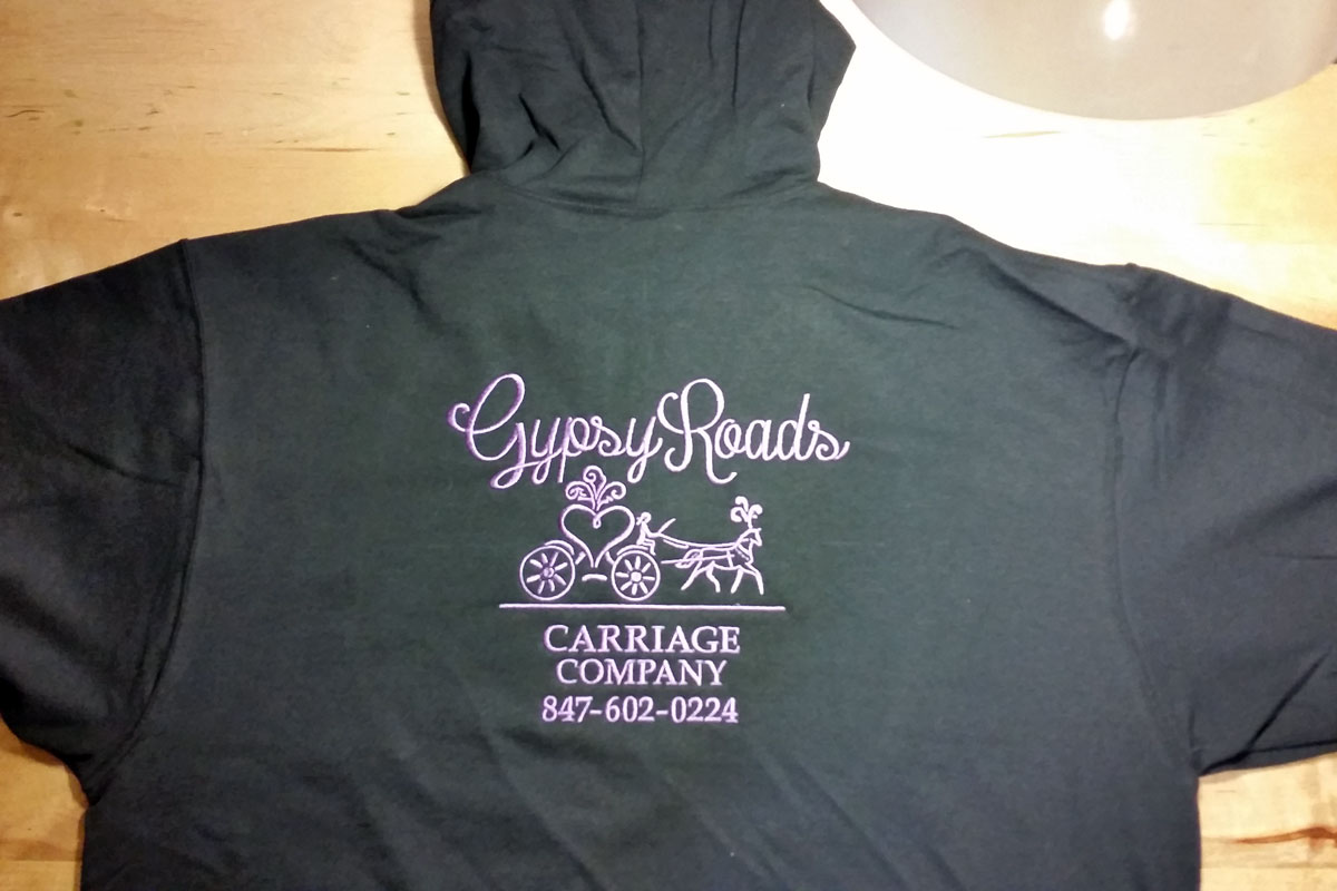 Black or dark gray hoodie with Gypsy Roads Carriage Company embroidered in light purple on the back with the image of a heart-shaped, horse-drawn carriage.