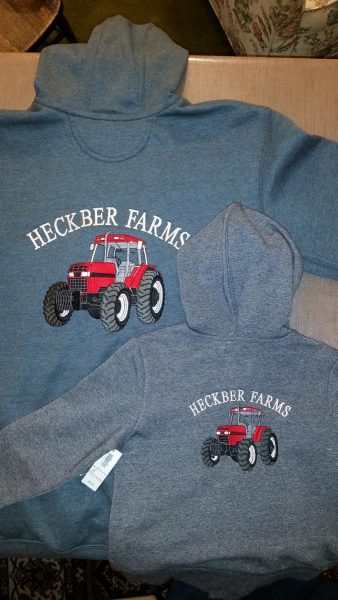 An adult-size gray hoodie laying face-down with a large red tractor embroidered on the back and the words Heckber Farms in white above the tractor. A youth size hoodie lays face-down beside the adult-size one, with matching embroidery on the back.