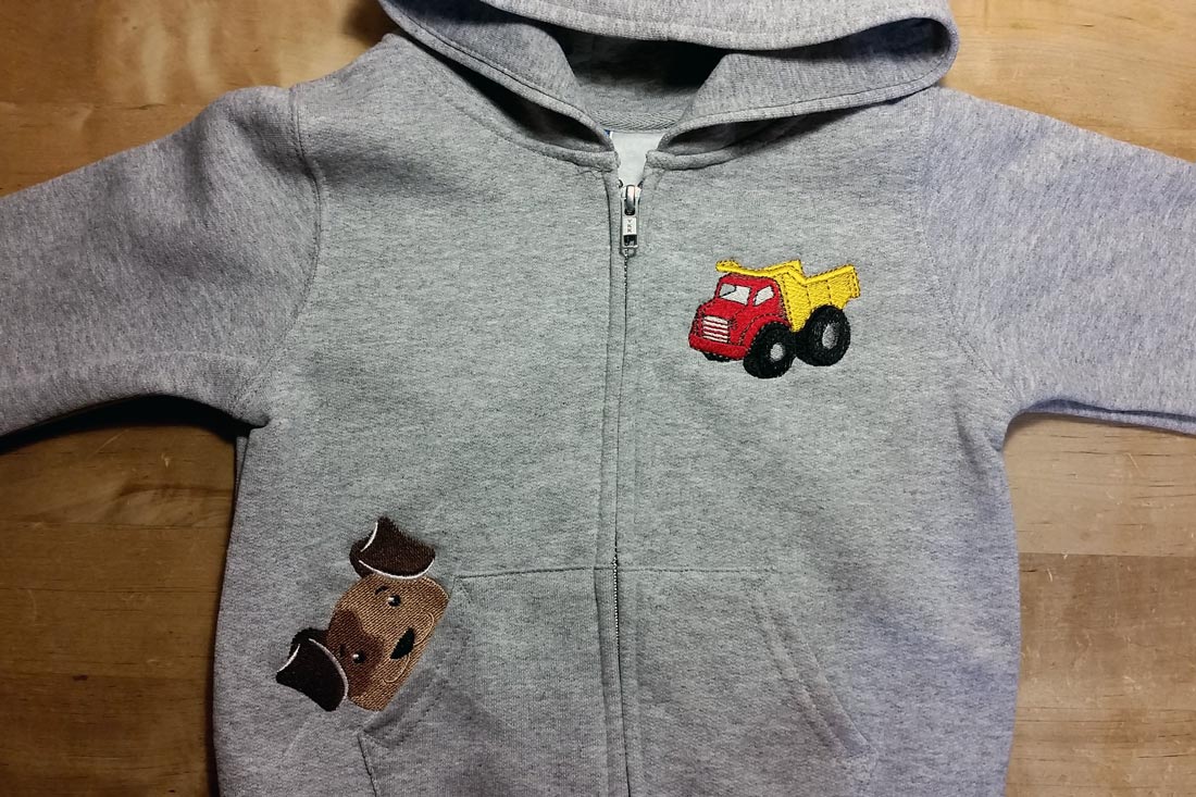A child's grey hoodie with an embroidered puppy dog peeking out of the right pocket and an embroidered dump truck on the left chest