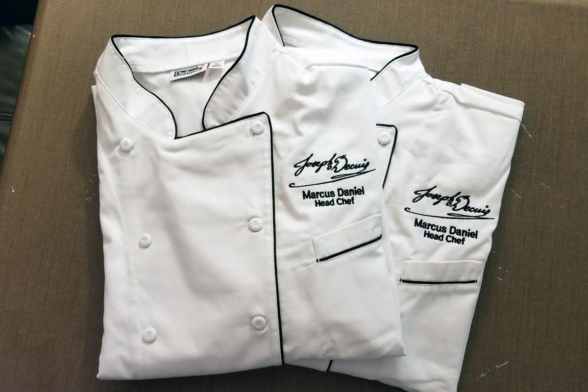 Two folded white chefs coats with black trim, each embroidered with the Joseph Decuis logo and Marcus Daniel Head Chef below the logo above the left chest pocket.