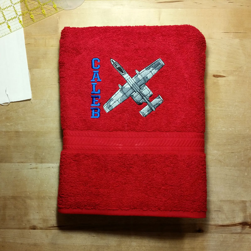 Child's red bath towel with an embroidered jet design and the name Caleb embroidered vertically in blue alongside the jet