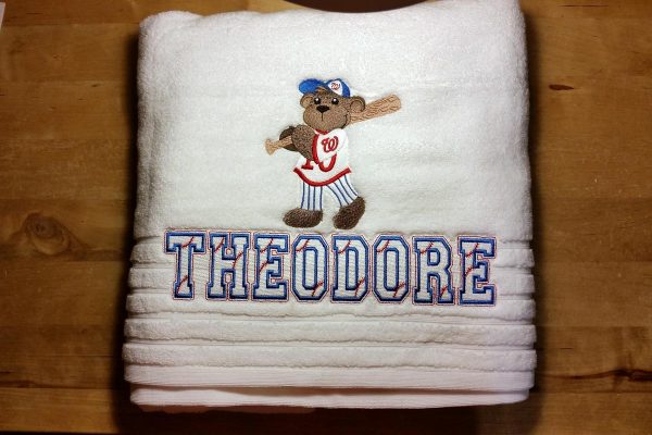 Child's white bath towel with a teddy bear baseball player carrying a bat embroidered above the name Theodore which is embroidered in a font that looks like it has baseball stitches running through it