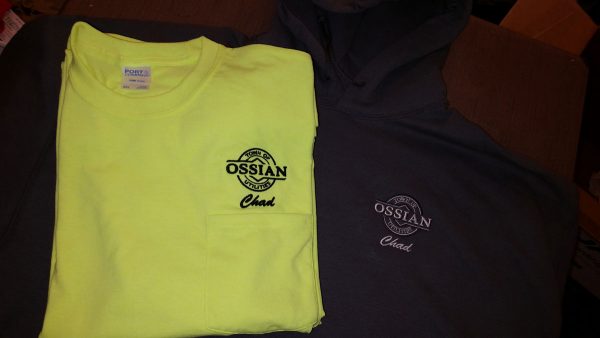 A folded, bright yellow t-shirt with the Town of Ossian Utilities logo and the name Chad embroidered above the left chest pocket sits on top of a dark gray hoodie with the same embroidery in silver.
