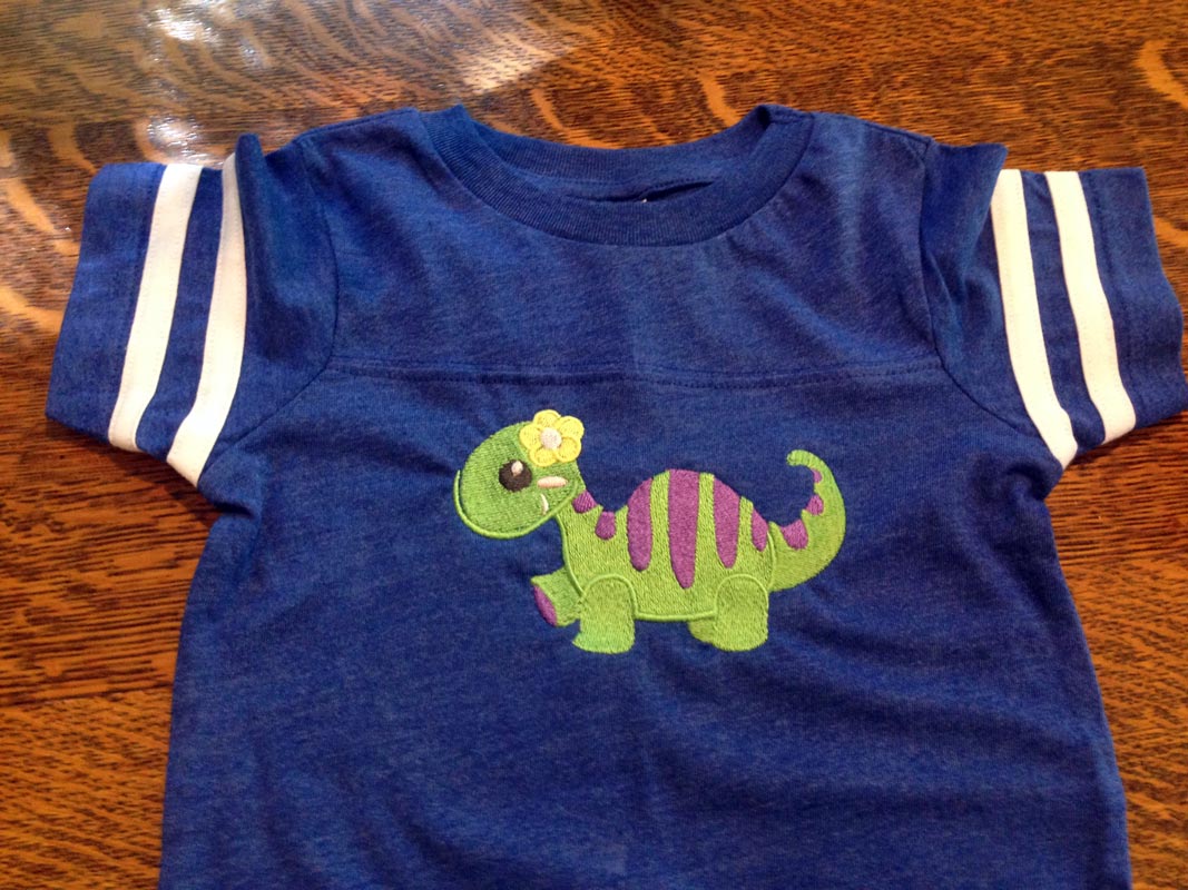 Child's blue t-shirt with an embroidered green youthful-looking dinosaur with purple stripes and a yellow flower on her head.