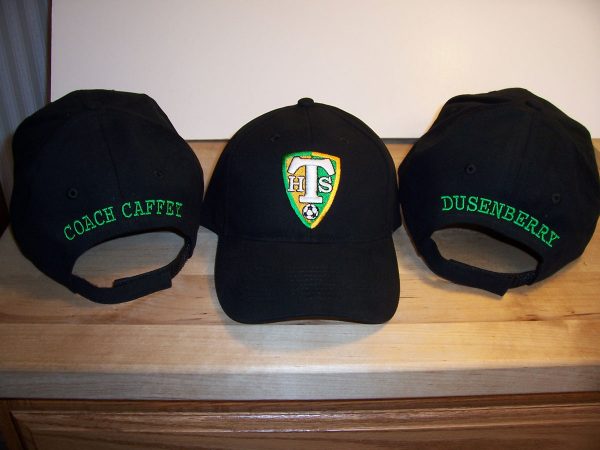Three black baseball caps. Two are facing backwards, one is facing forwards. One facing back is embroidered with Coach Caffey and the other with Dusenberry. The one facing the front is embroidered with a gold and green shield logo for Tumwater High School Soccer.